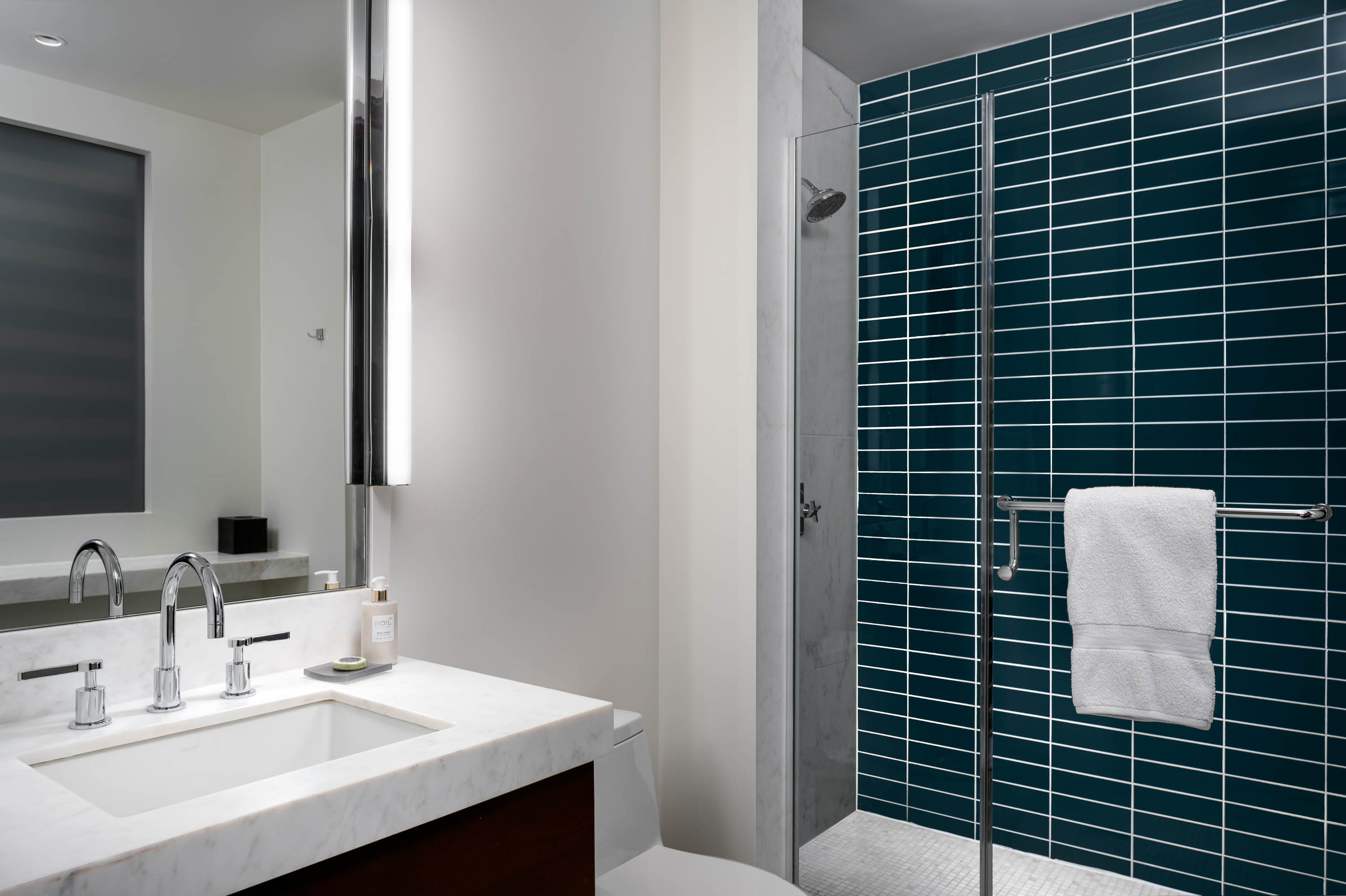 A modern bathroom with a white marble sink, dark wood accents, teal tile shower, and large mirror.