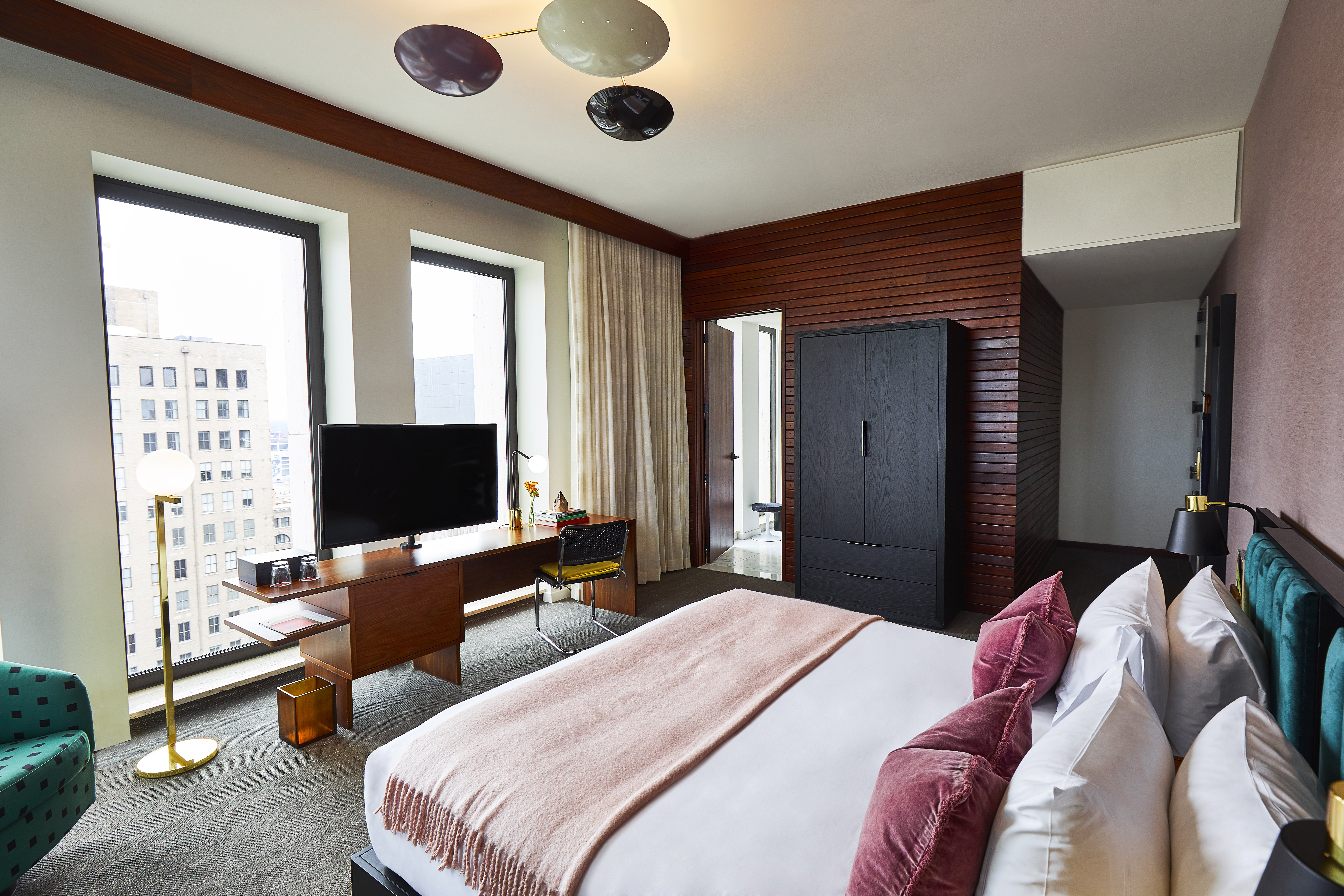 A hotel room with a king-sized bed, desk, TV, and wardrobe, overlooking the city.